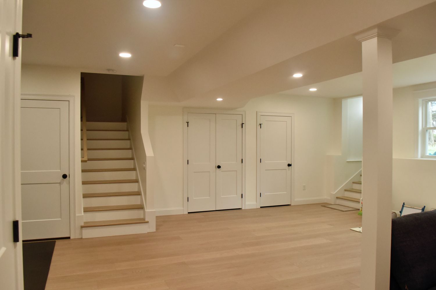 A spacious, well-lit basement with beige walls, light wood flooring, and white double doors.