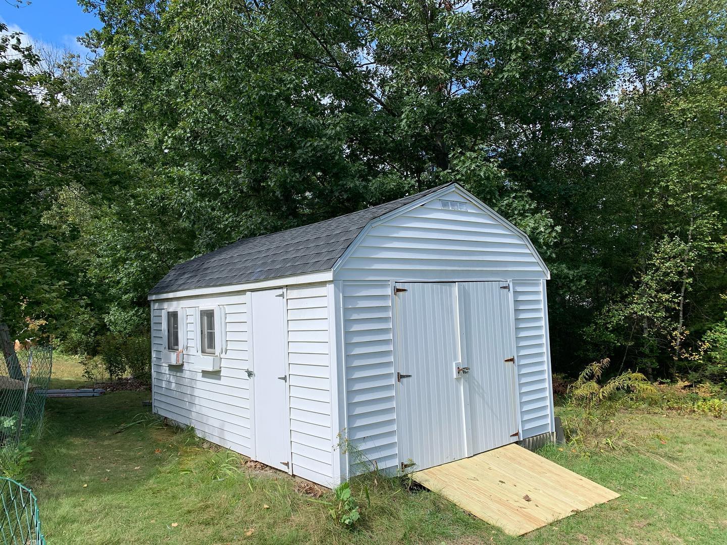 White garden shed with double doors and small windows in a grassy backyard.