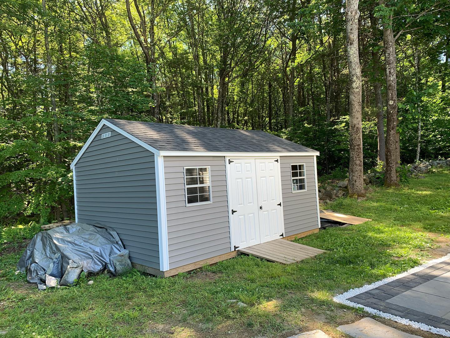 A gray garden shed with white doors in a wooded backyard.